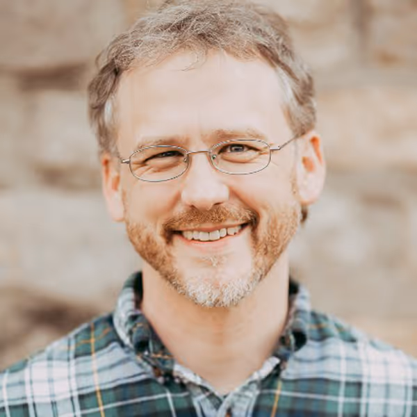 Smiling middle-aged man with short curly hair and beard wearing glasses and a green plaid shirt.