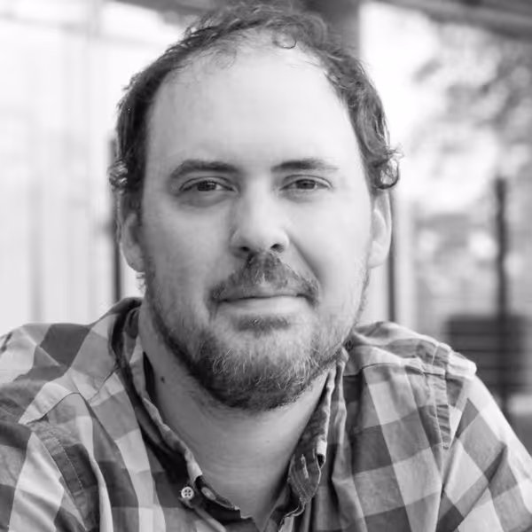 Black-and-white portrait of a man with short curly hair and a beard, wearing a checkered shirt and looking directly at the camera.