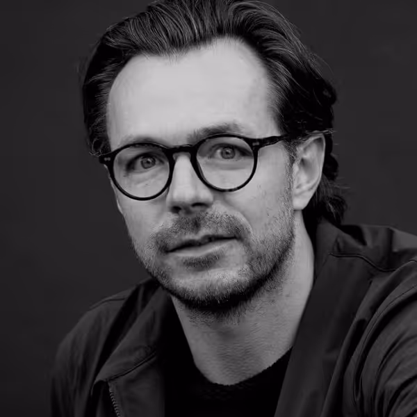 Black and white portrait of a man with short hair, stubble, and round glasses, wearing a jacket over a shirt.