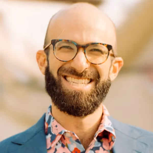 Smiling bald man with beard wearing glasses, a colorful floral shirt, and a blue blazer.