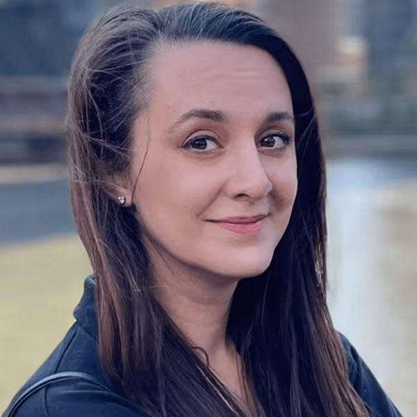Smiling woman with long brown hair wearing a dark jacket standing outdoors near a body of water.