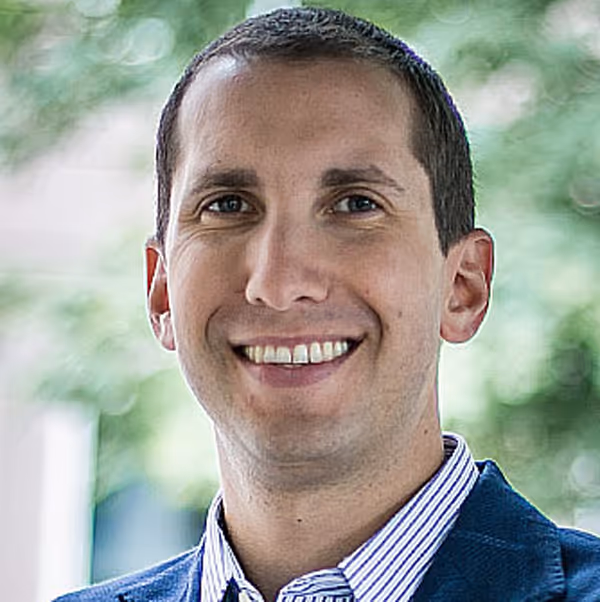Smiling man with short hair wearing a blue suit jacket and striped shirt, standing outdoors with blurred green background.