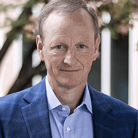 Smiling middle-aged man wearing a blue checkered blazer and light blue shirt, standing outdoors with blurred trees in the background.