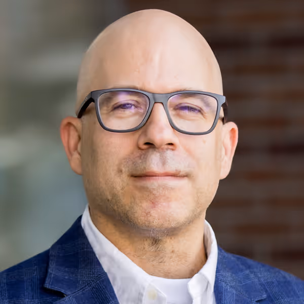 Portrait of a bald man wearing glasses, a white shirt, and a blue blazer with a blurred background.