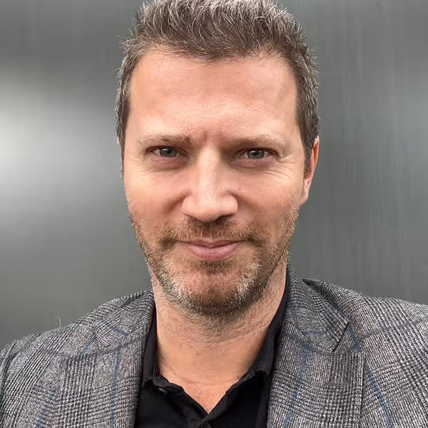 Close-up portrait of a man with short hair and a trimmed beard wearing a checked blazer and black shirt against a plain gray background.