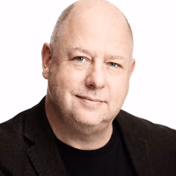 Portrait of a middle-aged bald man with light skin, blue eyes, and slight stubble, wearing a black blazer and black shirt, smiling gently against a white background.