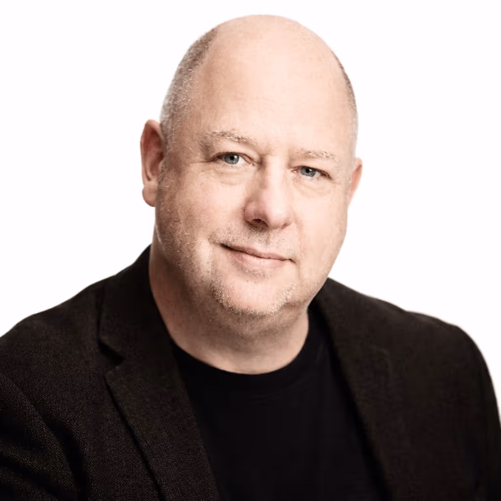 Middle-aged bald man wearing a black blazer and shirt, smiling gently against a white background.