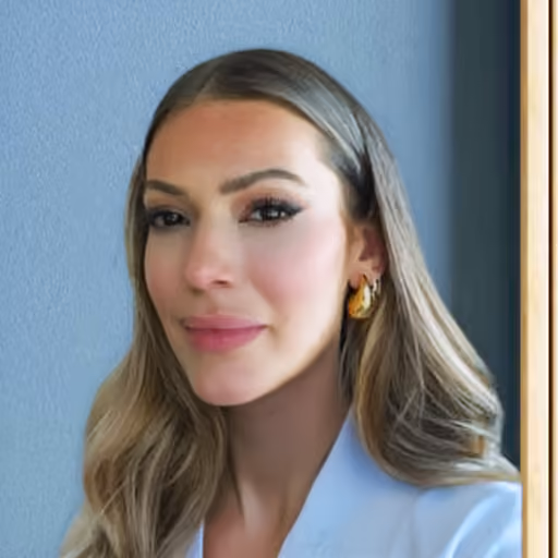 Smiling woman with long hair wearing gold earrings and a light blue blazer against a blue wall background.