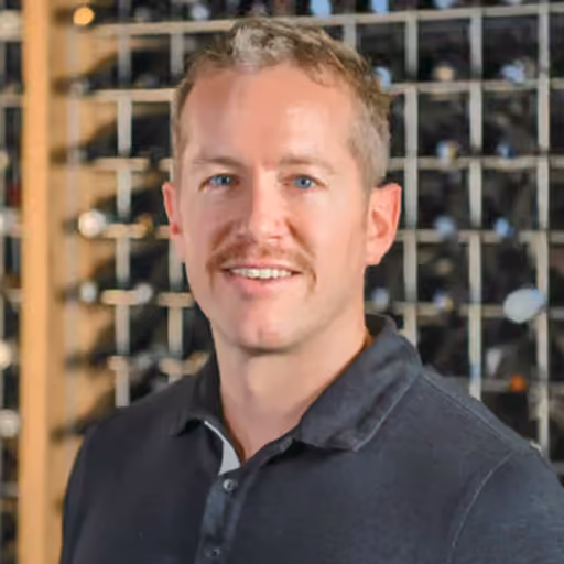Man with short blond hair and mustache wearing a dark polo shirt in front of wine bottles on shelves.
