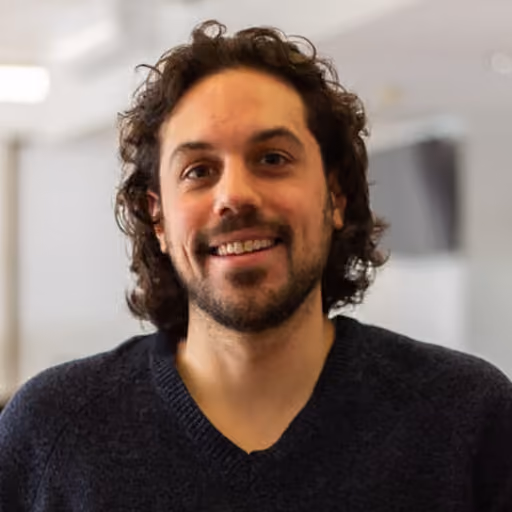 Smiling man with curly dark hair and beard wearing a dark sweater in a blurred indoor setting.