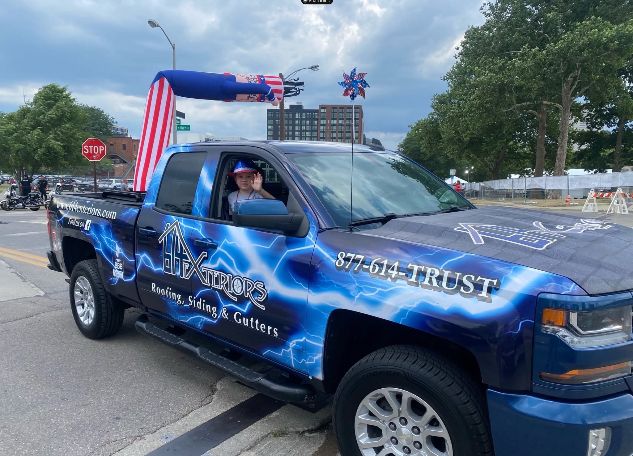 Child waving from a blue 614 Exteriors truck decorated for a patriotic community parade.