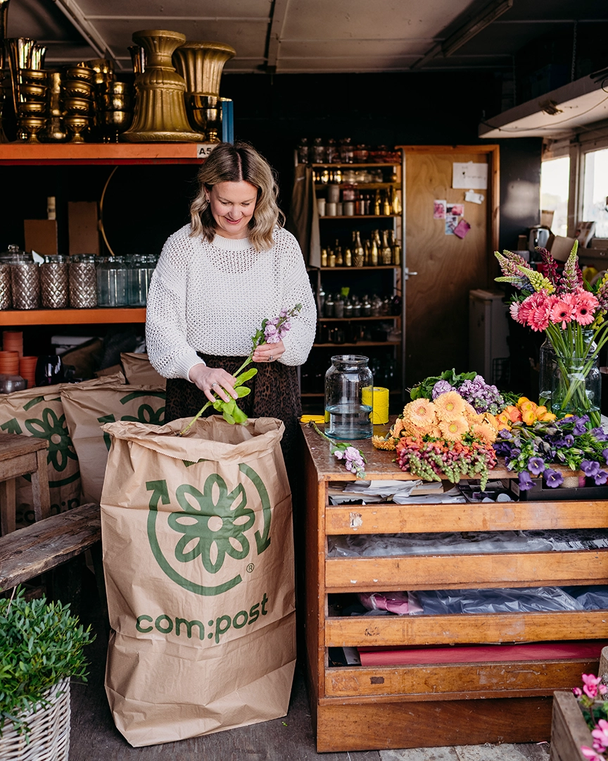 A woman in a white sweater placing flowers into a large brown compost bag in a workshop filled with flowers, glass jars, and shelves with vases and supplies.