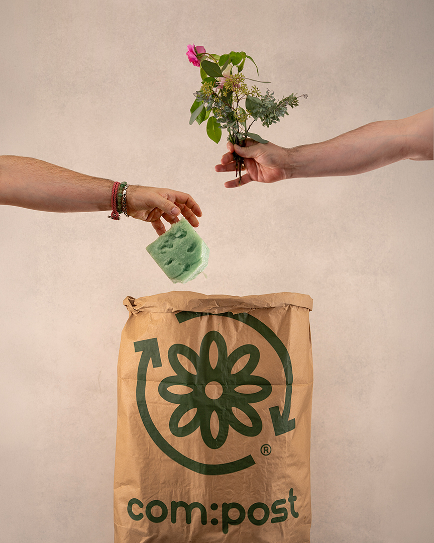 Two hands above a brown compost bag with a green flower logo and arrows forming a circle; one hand holds a small bouquet of flowers and leaves, the other holds a green floral foam, both positioned as if about to be placed into the compost bag.