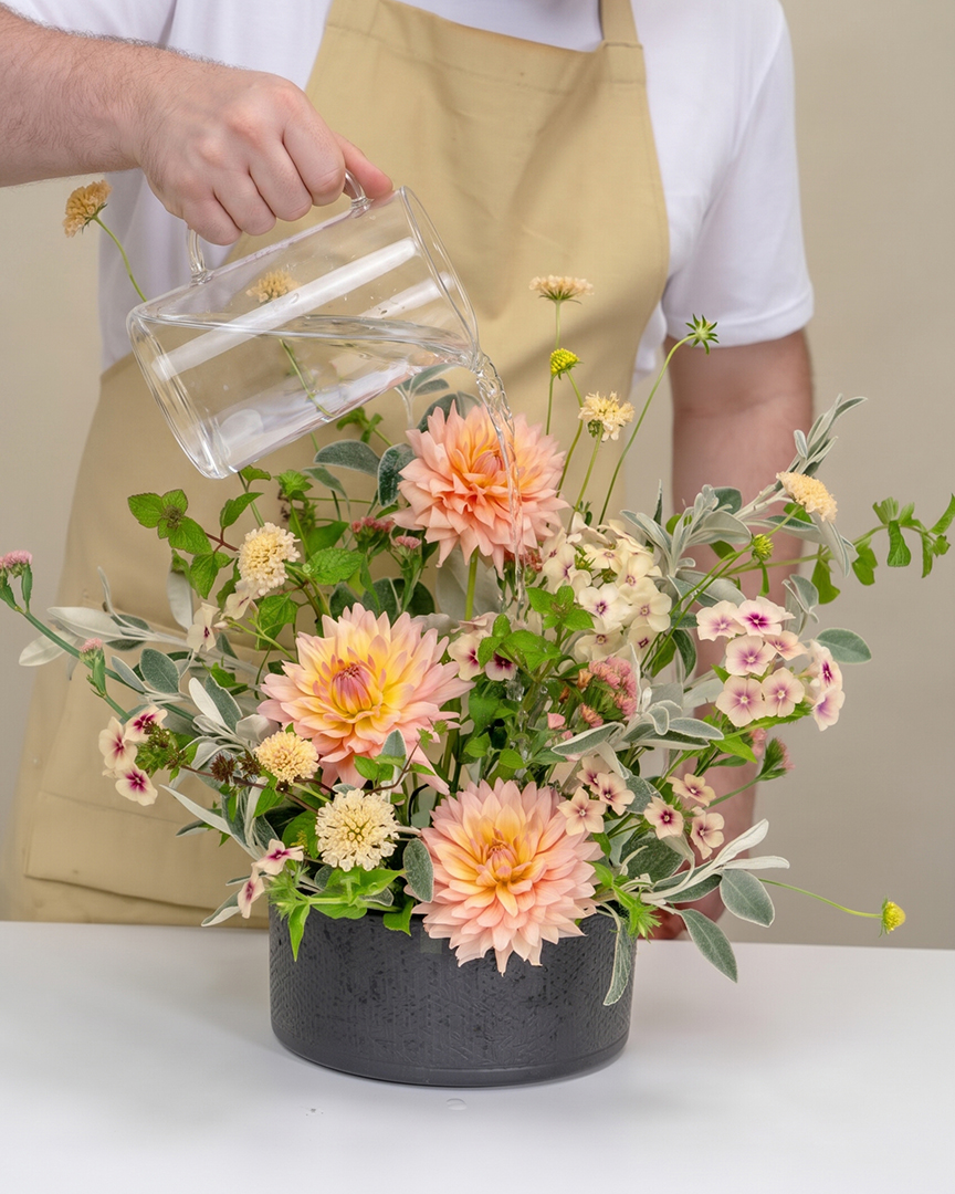 Person wearing a beige apron and white shirt watering a floral arrangement of peach dahlias and assorted green and white foliage in a black round container with a clear glass pitcher filled with water.