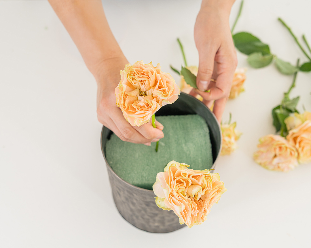 Person arranging peach-colored roses into green floral foam inside a black hammered metal container, with more roses and leaves scattered on a white surface.