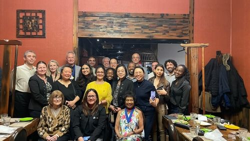 A group of about 20 diverse people looking at the camera and smiling in a orange room with coats hanging on the right side and two dinner tables. Most of the people are standing, only 3 are sitted.
