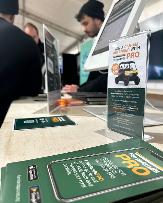 Trade show booth display featuring Farmlands promotional materials on a wooden table, including green brochures with 'PRO' branding and a standing banner advertising a Can-Am Defender Pro giveaway contest, with people in business attire visible in the background