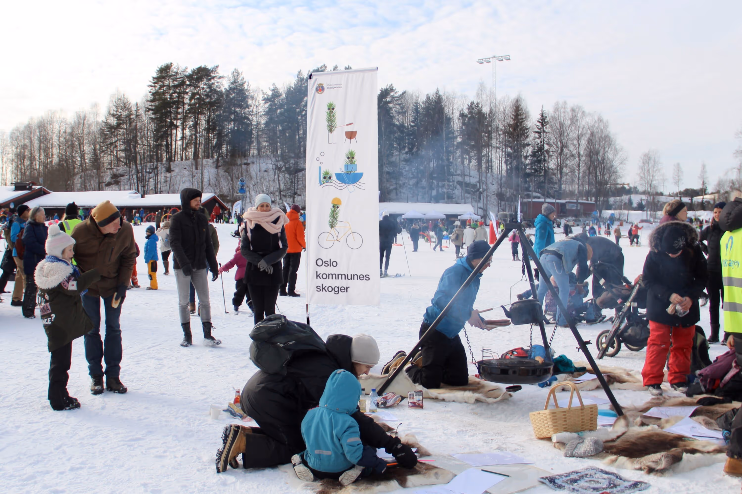 People gathered on snow-covered ground around a campfire cooking setup and engaging in outdoor activities near a banner that reads 'Oslo kommunes skoger.'