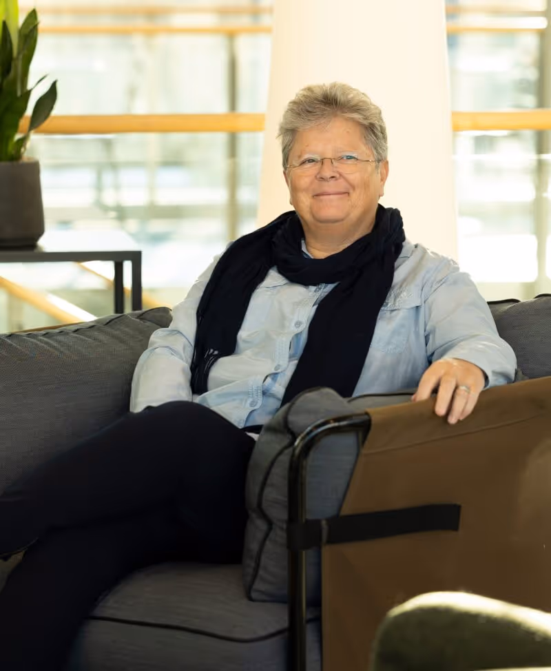 Gerd smiling. She has short hair and glasses sitting on a sofa wearing a light blue shirt and black scarf.