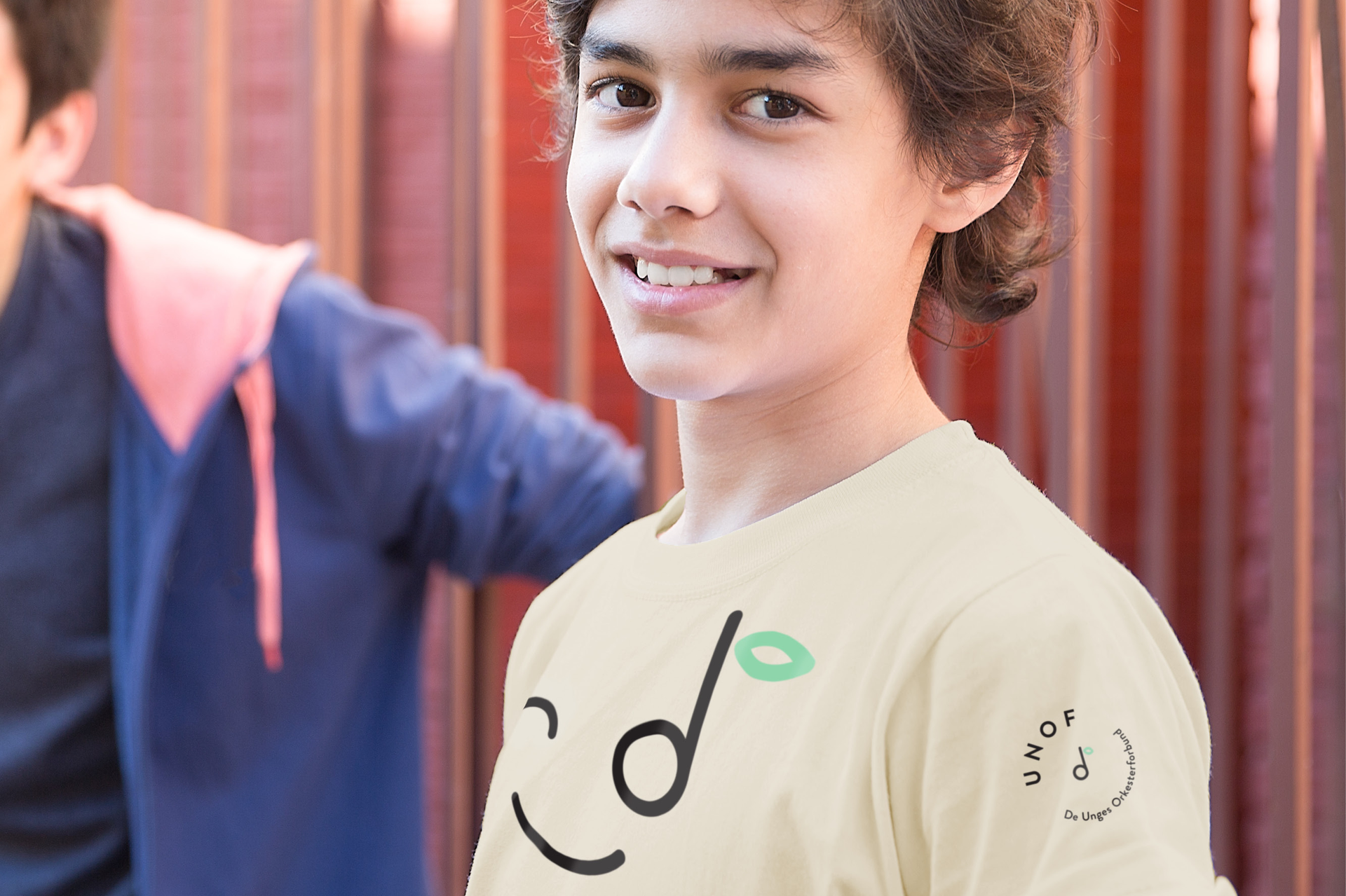 Smiling teenage boy wearing a beige t-shirt with a musical note and a green leaf design, standing in front of a blurred person in a blue jacket.