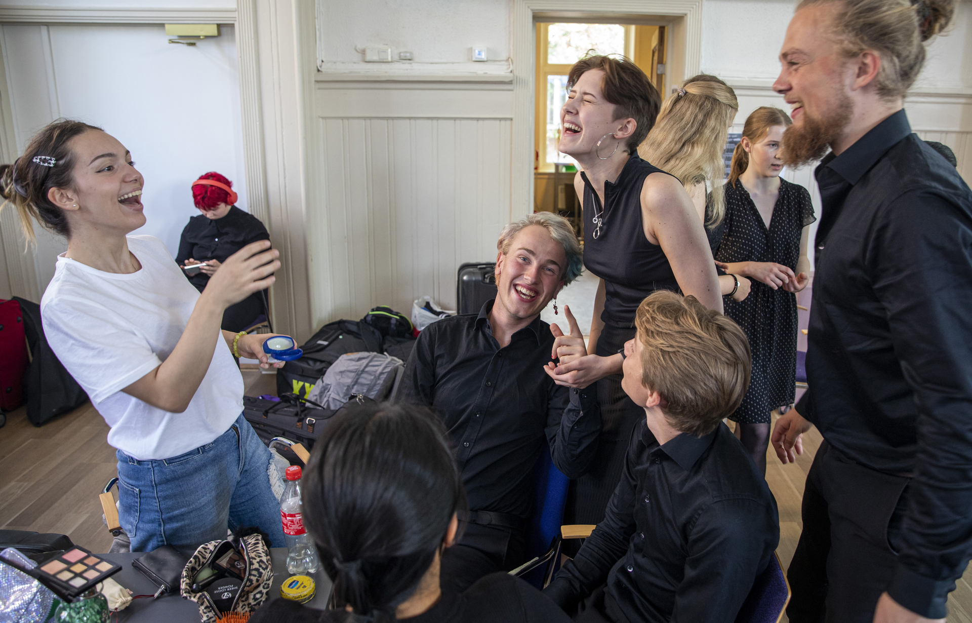Group of young people laughing and socializing indoors, some sitting and some standing near a table with makeup and a drink bottle.