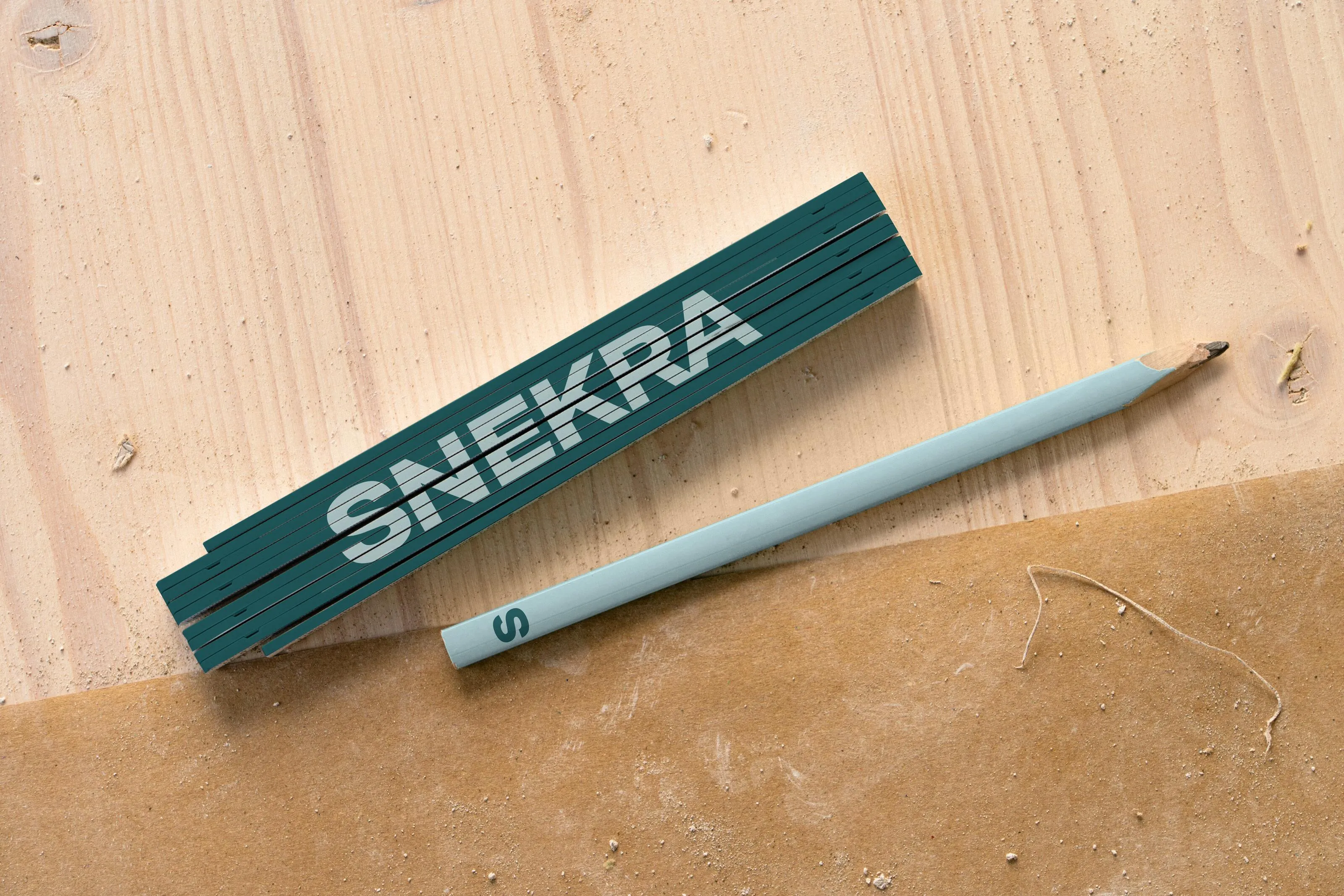 Green folding wooden ruler labeled SNEKRA beside a light blue pencil on wood and cardboard surfaces.