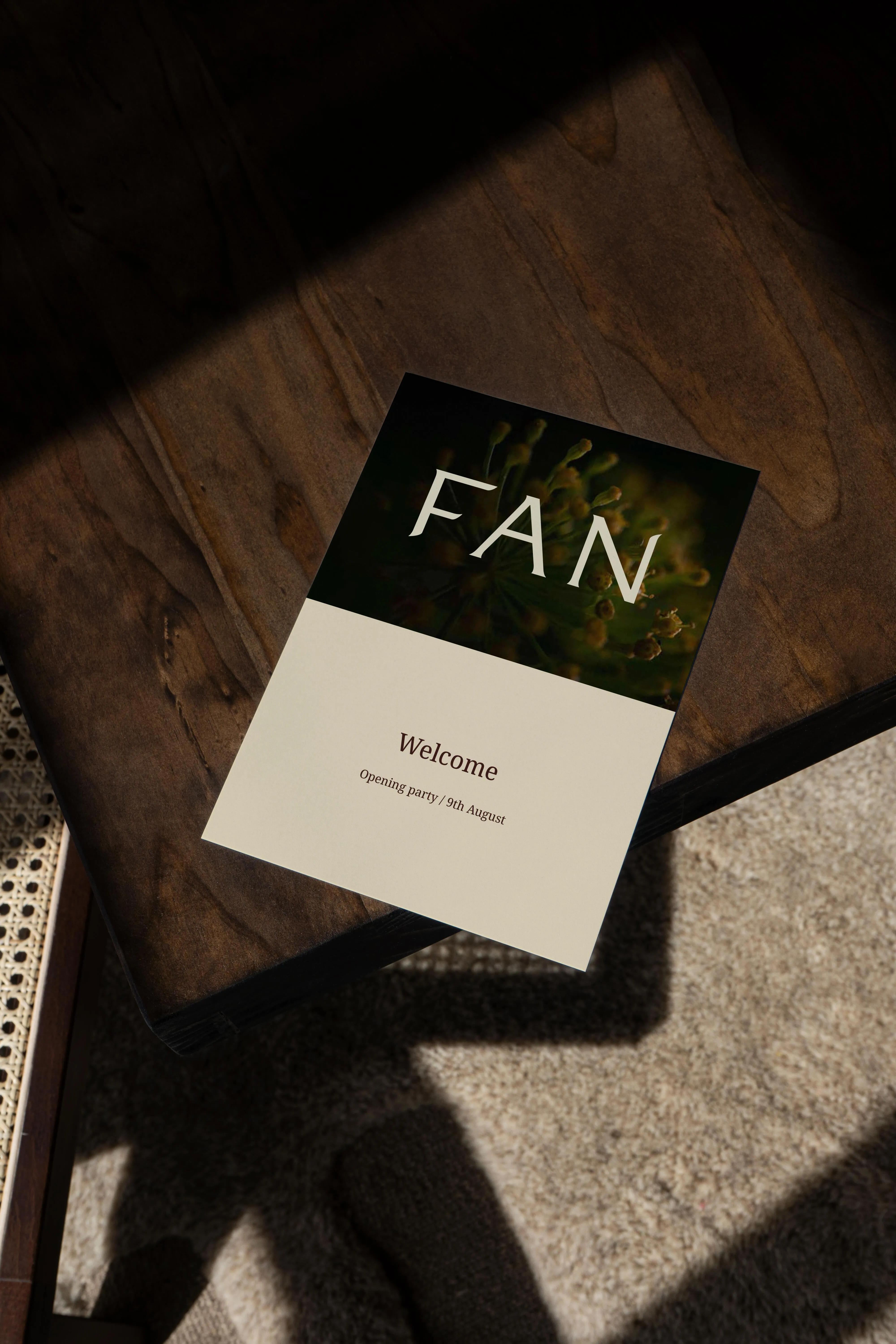 Invitation card with text 'FAN' and 'Welcome Opening party / 9th August' placed on a wooden table with shadows.