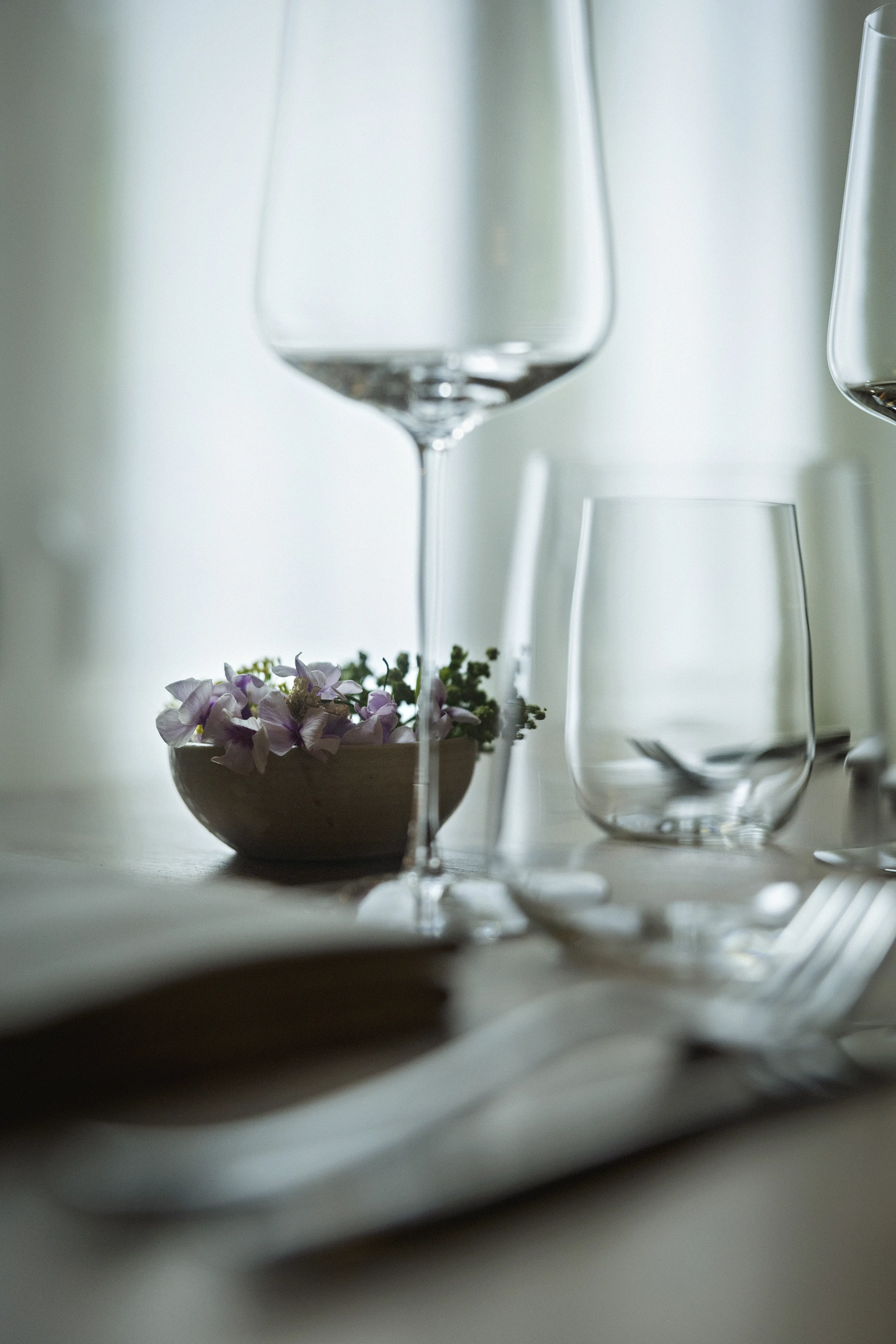 Close-up of a dining table with empty wine and water glasses, silverware, and a small bowl of purple flowers and greenery.