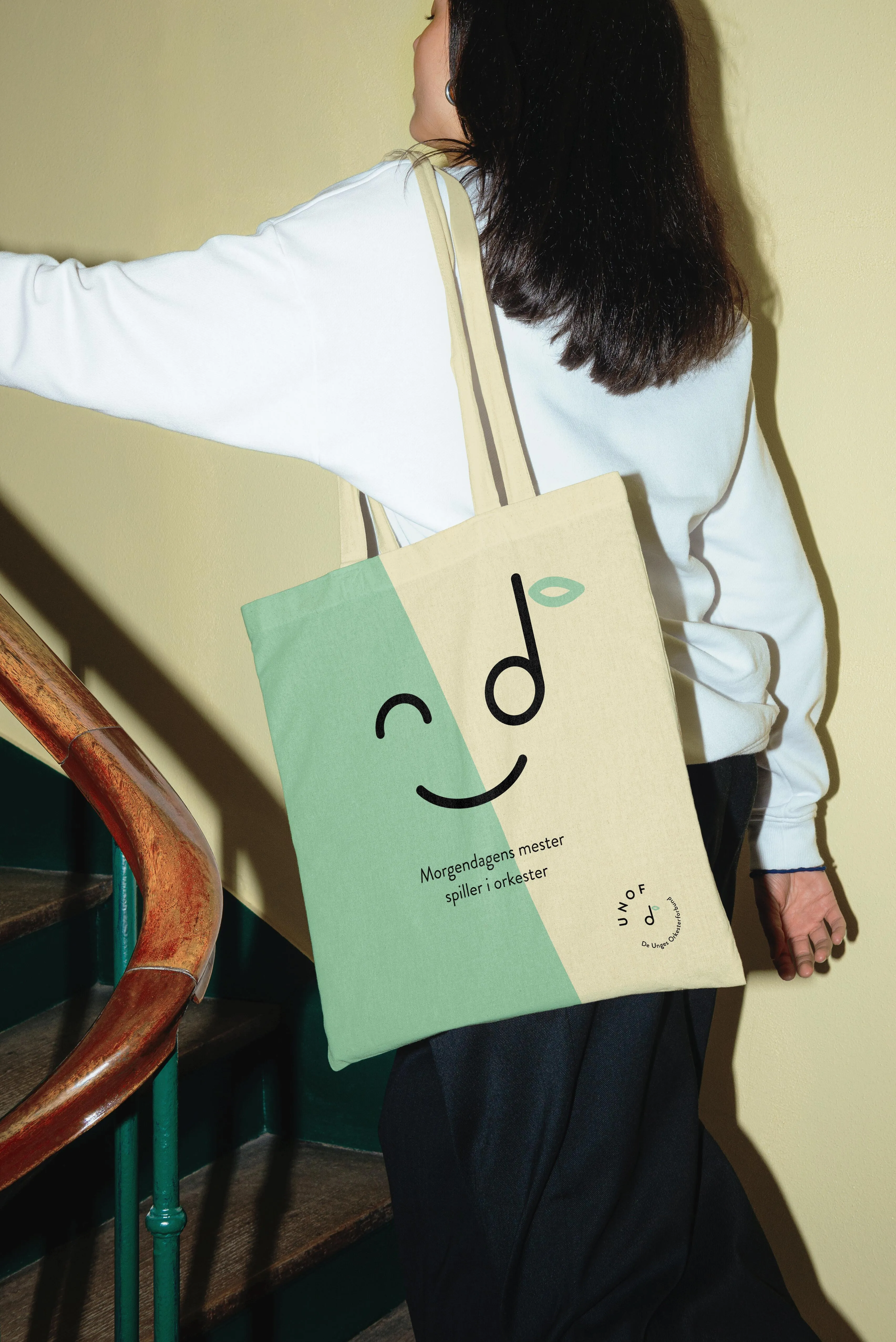Person with shoulder-length dark hair wearing a white sweater and carrying a beige and green UNOF tote bag with a smiling face design and text in Norwegian.