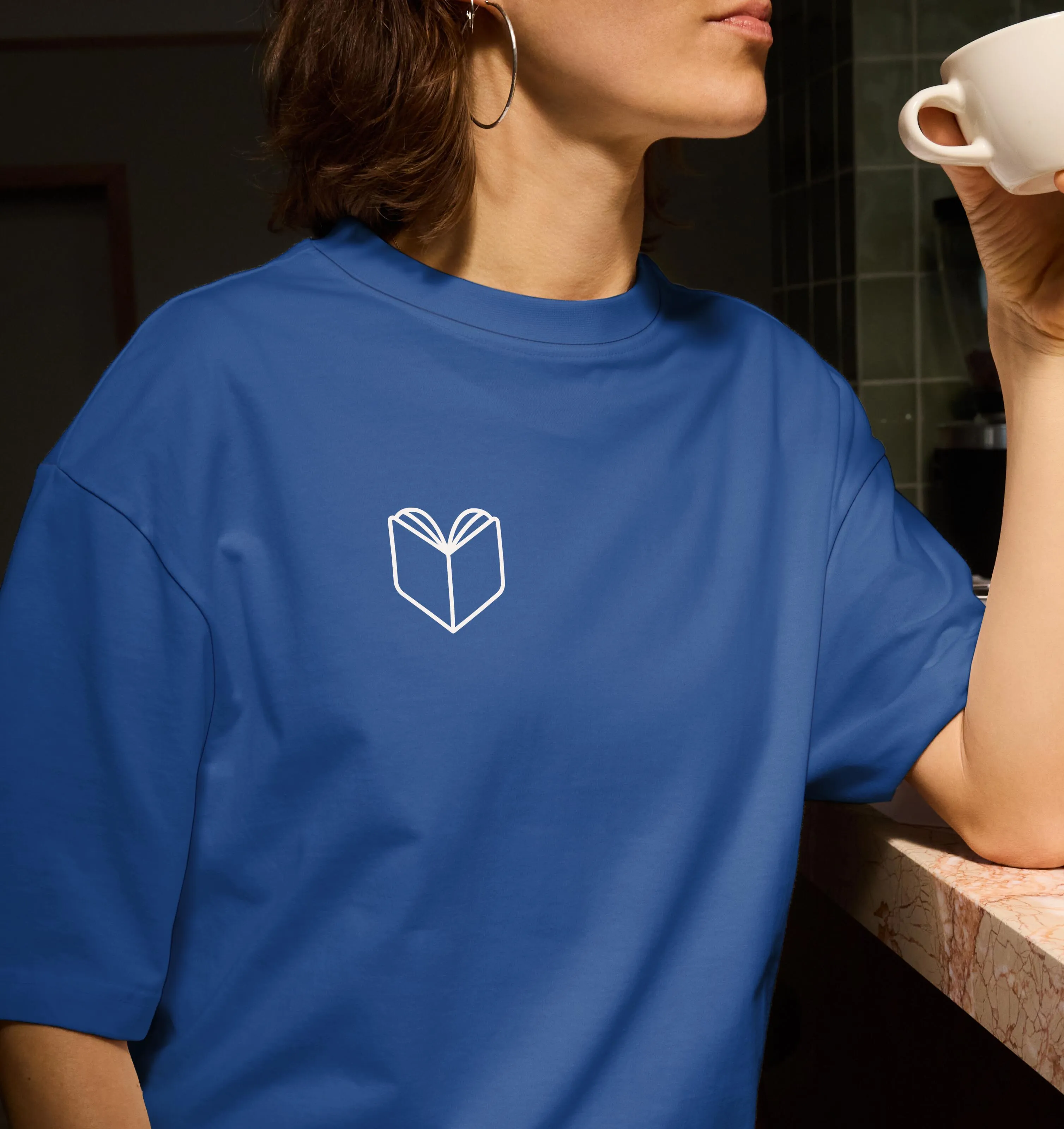 Person wearing a blue t-shirt with a white minimalist open book icon on the chest, holding a white cup near their mouth.
