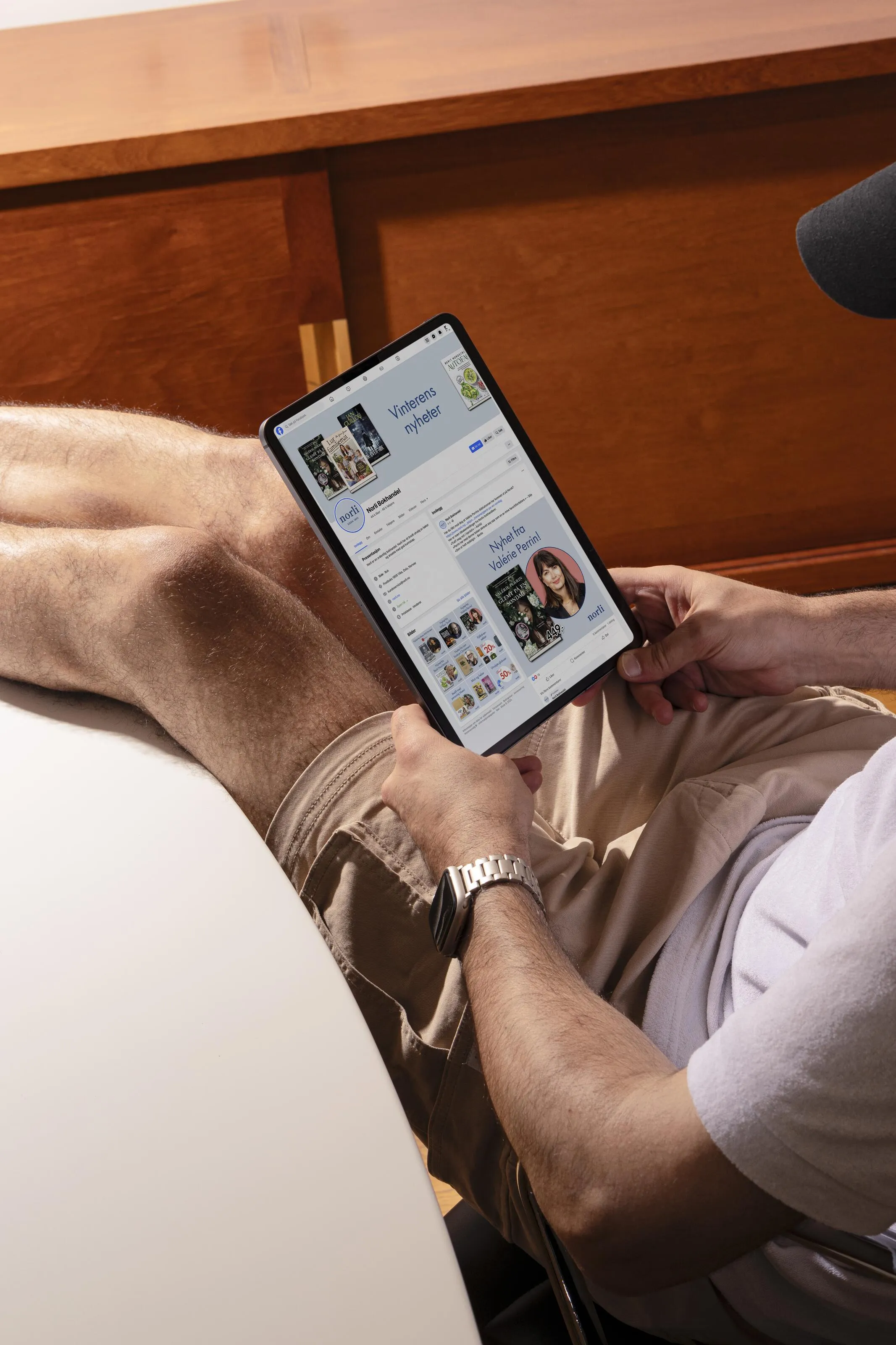 Person sitting with legs crossed, holding a tablet displaying Norli's Facebook page.