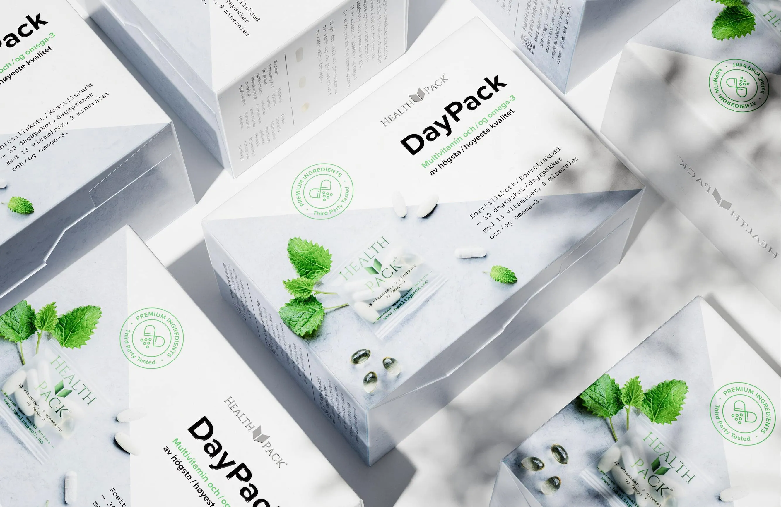 Boxes of Health Pack DayPack multivitamins with omega-3 capsules and fresh green leaves on a light surface.