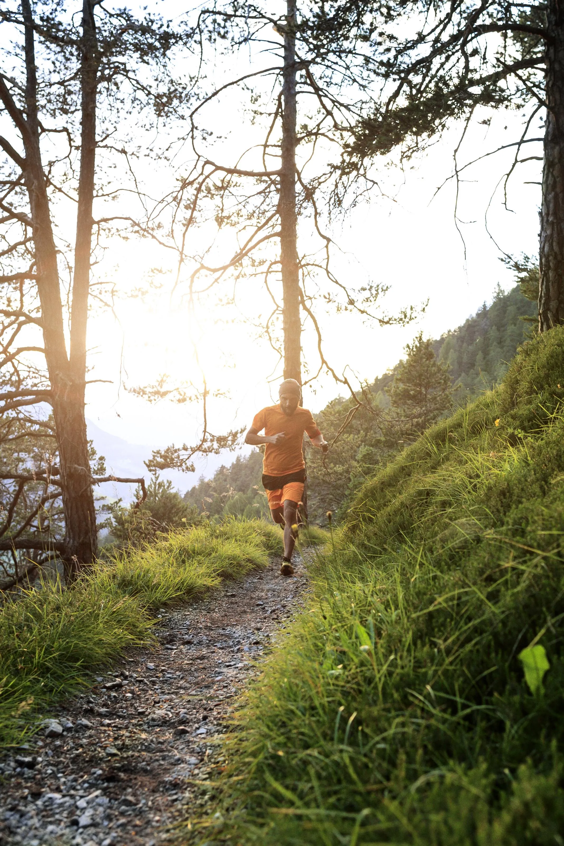 Man wearing orange running on a dirt trail through a forested area with sunlight filtering through trees.