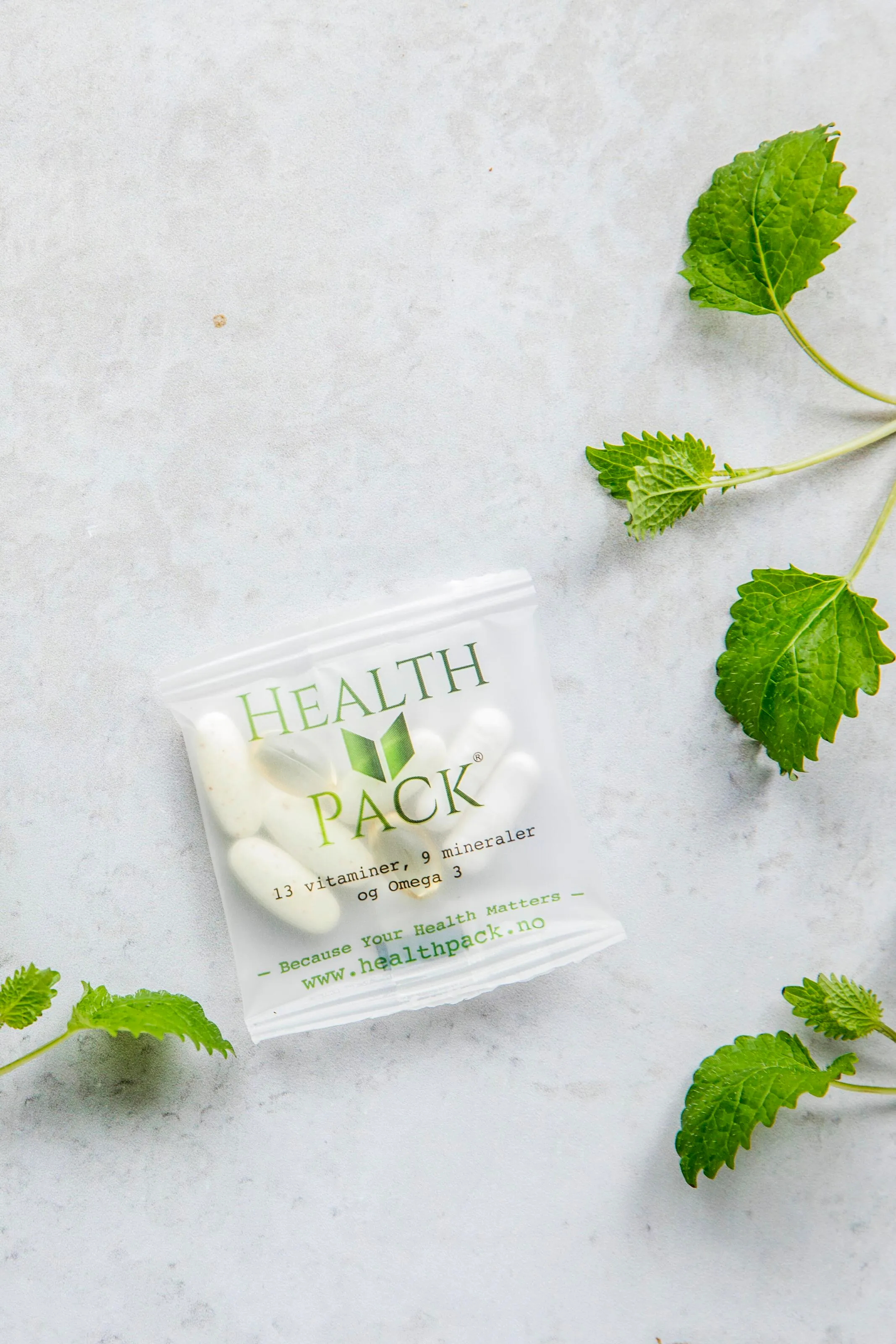 Plastic packet labeled Health Pack containing white supplement capsules on a light surface with green leaves around.