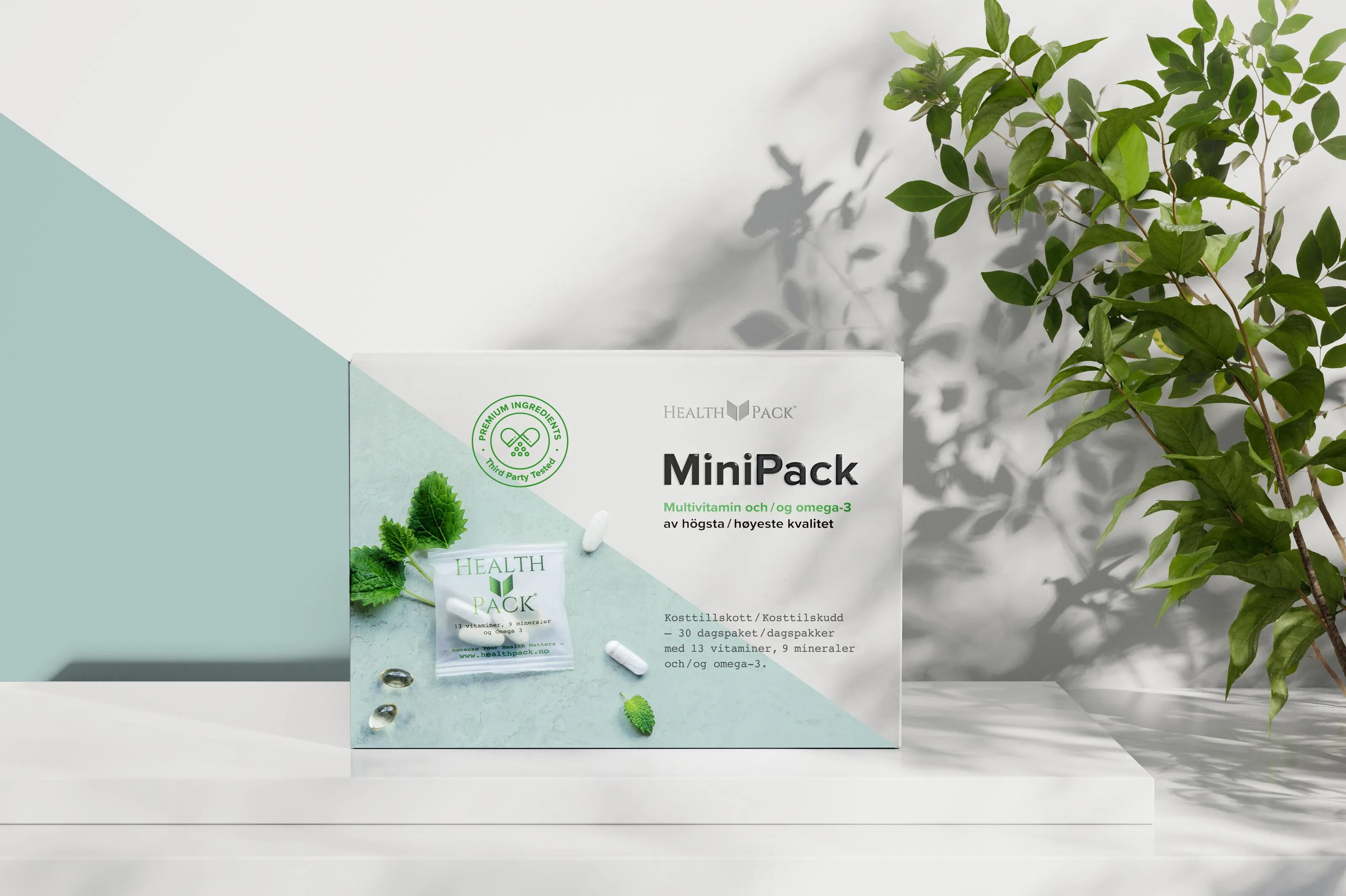 Box of Health Pack MiniPack multivitamin and omega-3 supplements with pills and green leaves, placed on a white surface with a plant shadow in the background.