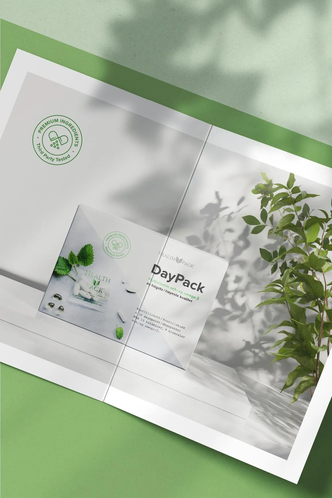 Open brochure showing a Health Pack DayPack multivitamin and omega-3 supplement box next to a green leafy plant, with shadows on a green and white background.