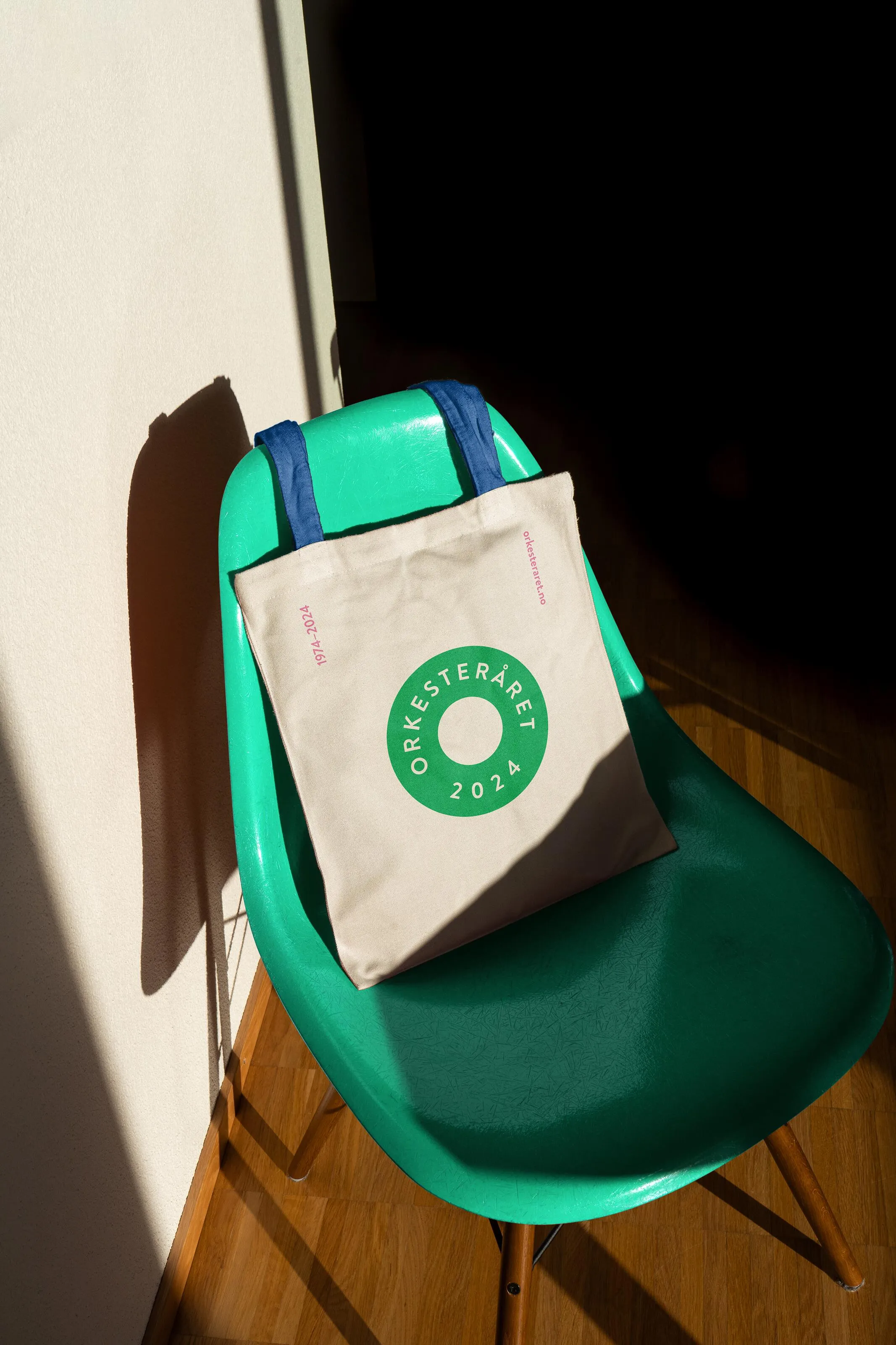 Canvas tote bag with green Orkesteråret 2024 circle logo hanging on a bright turquoise chair in sunlight, demonstrating everyday use of the festival branding.