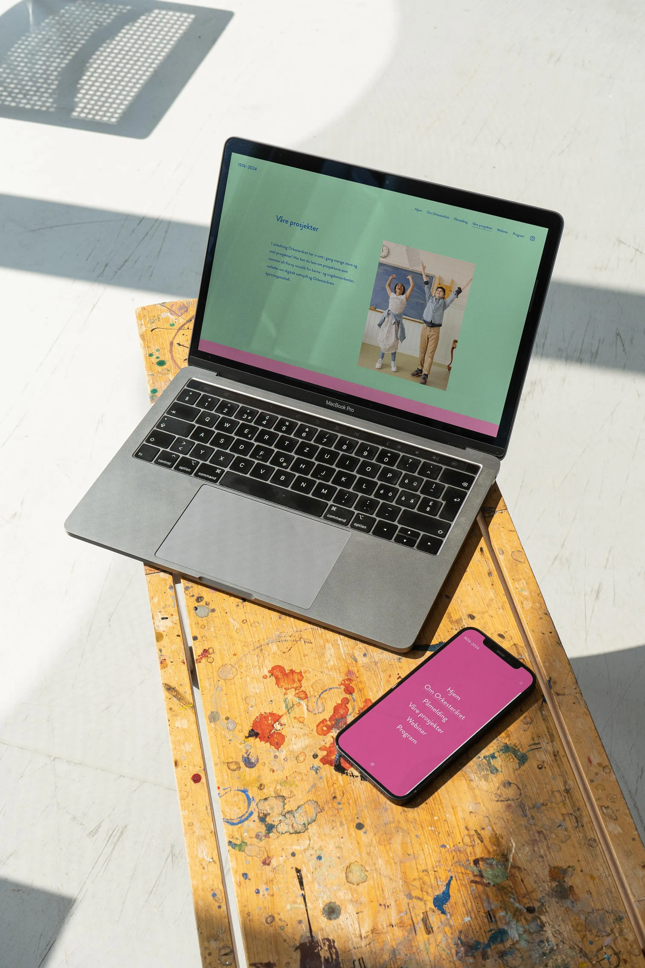 Open laptop and smartphone on a paint-splattered school table showing the Orkesteråret website and menu, demonstrating the colourful festival branding across digital devices.