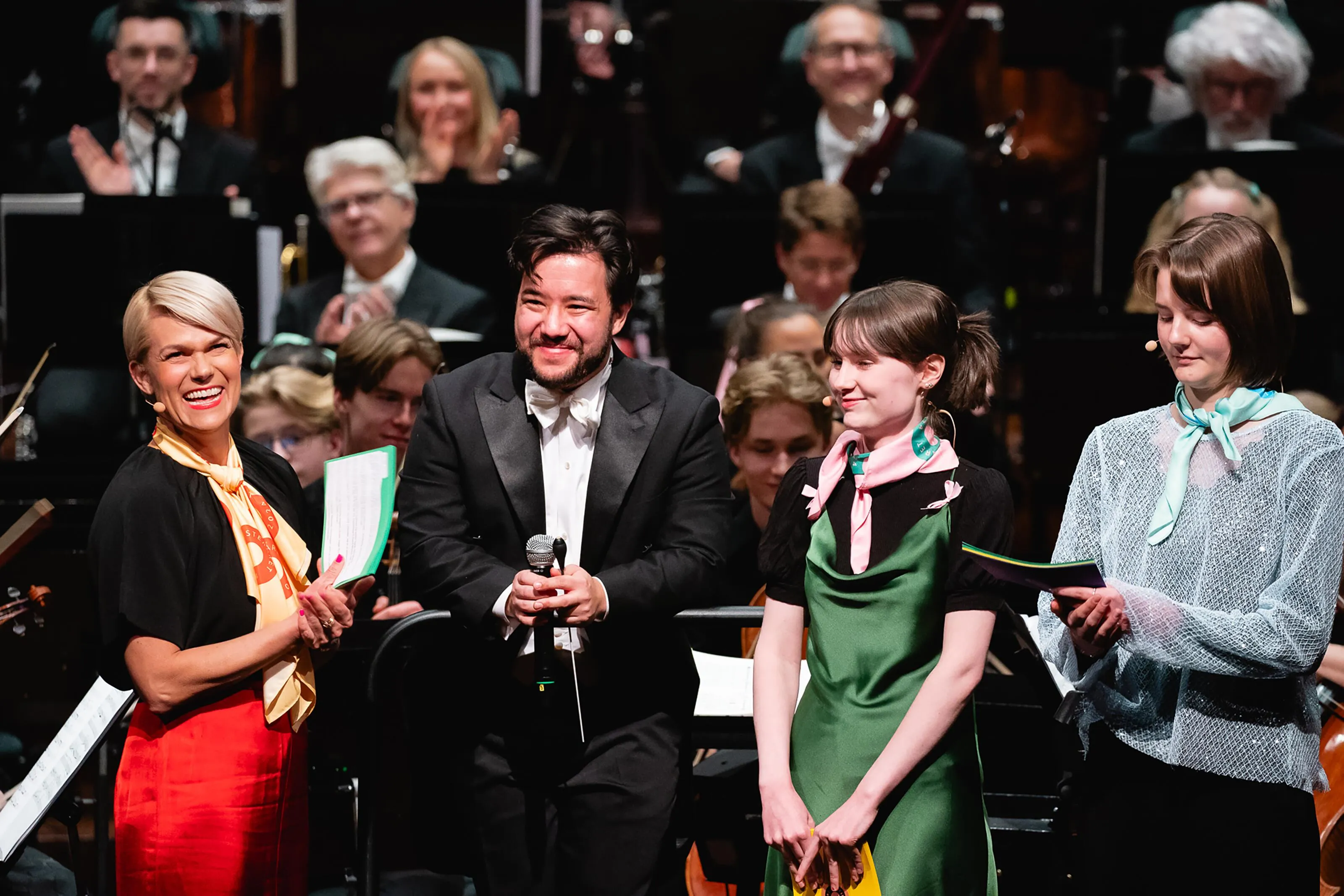 Four smiling presenters standing in front of a full symphony orchestra during an Orkesteråret 2024 concert, highlighting collaboration between professionals and young musicians.