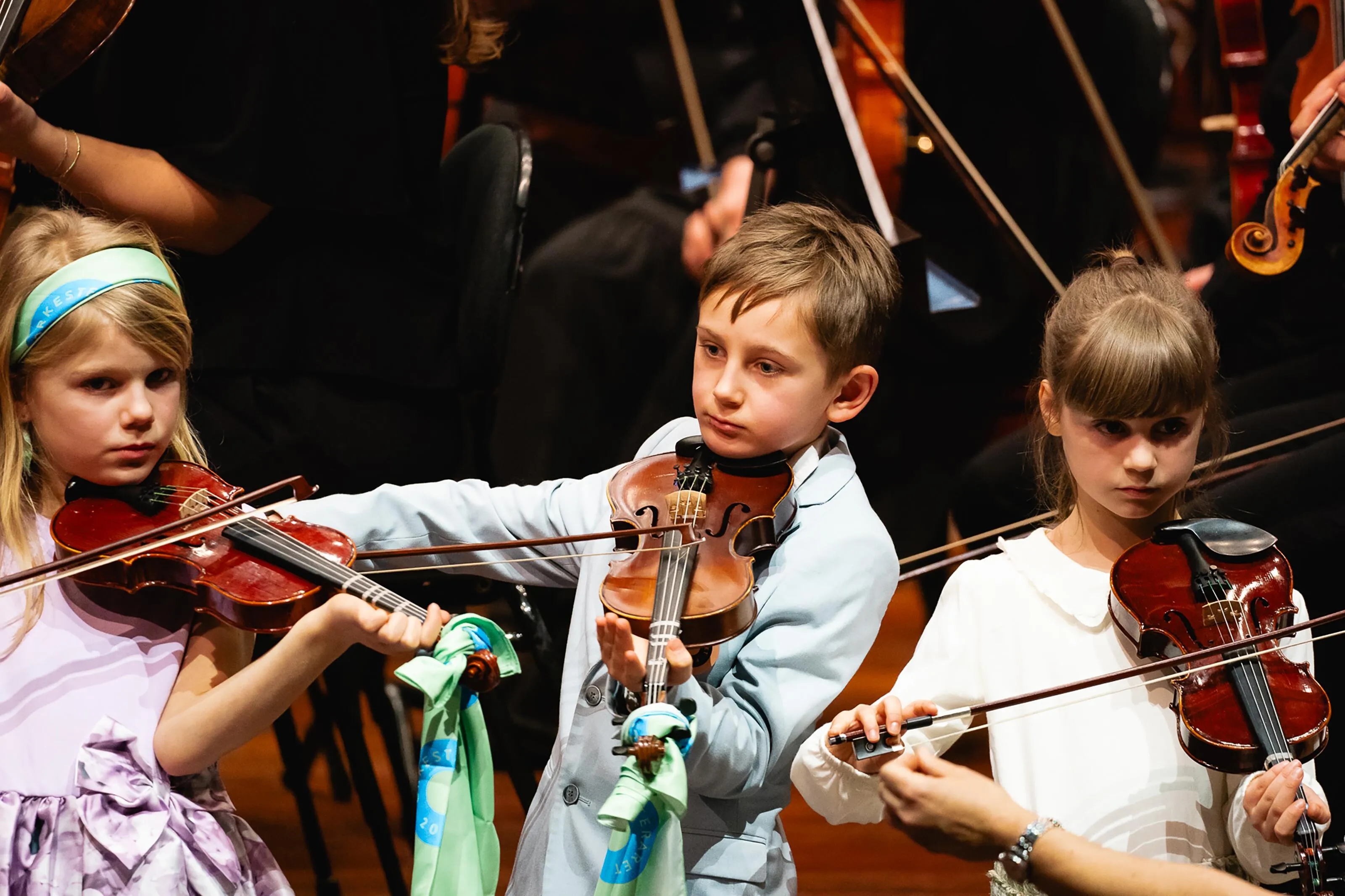 Three young children playing violins on stage during an Orkesteråret concert, each wearing colourful accessories, illustrating the project's focus on inclusive music-making for kids.