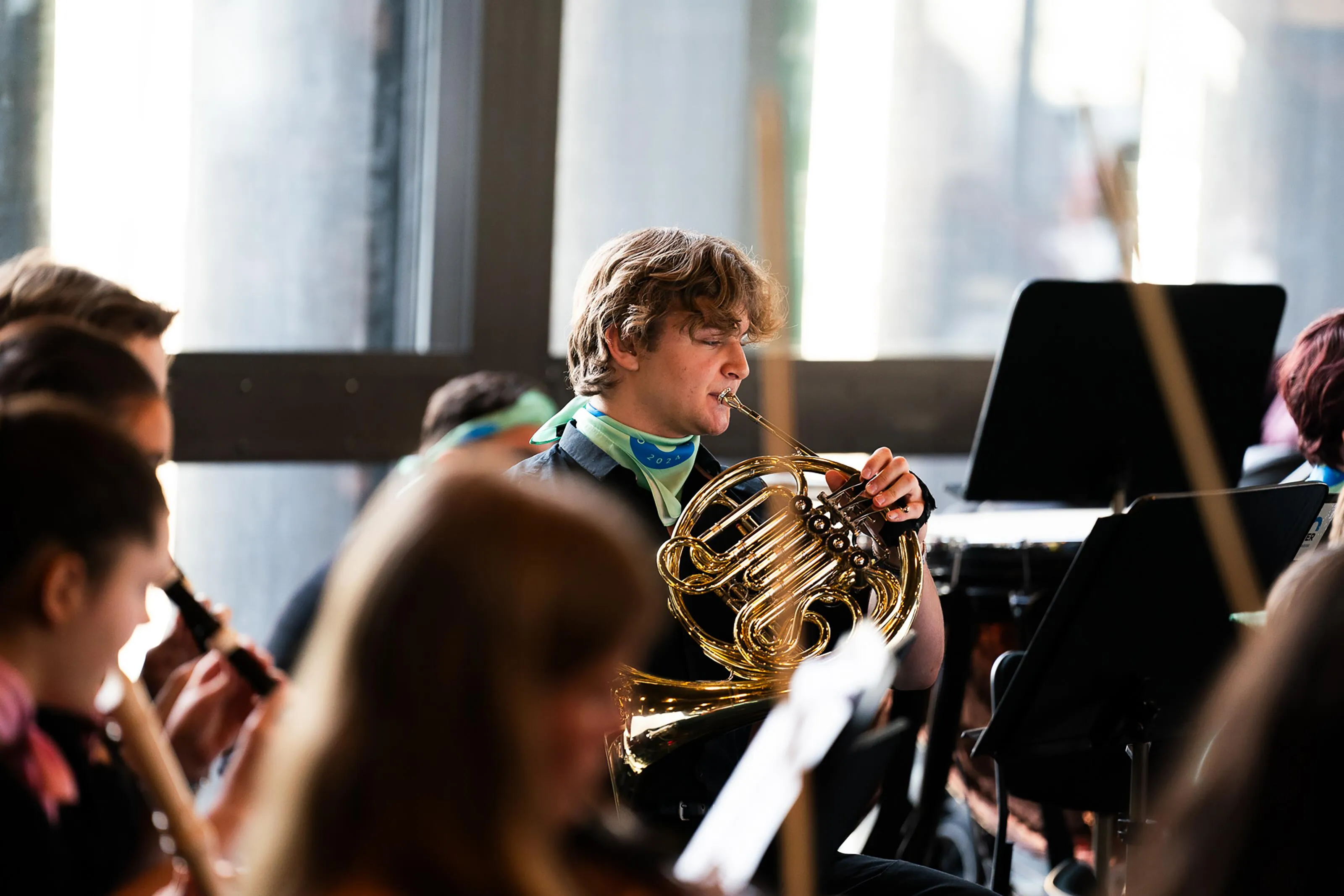 Young French horn player wearing an Orkesteråret scarf performing with a youth orchestra in a bright concert hall, surrounded by fellow musicians and music stands.