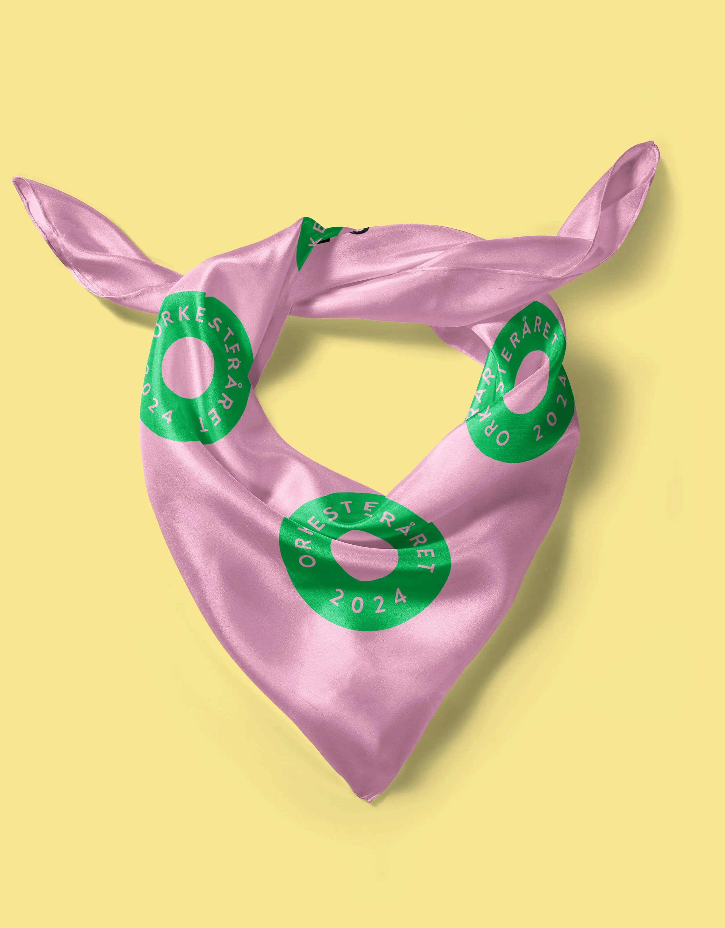 Pink Orkesteråret 2024 scarf laid on yellow background, printed with repeating green circle logos, showing how the visual identity is applied to wearable merchandise.