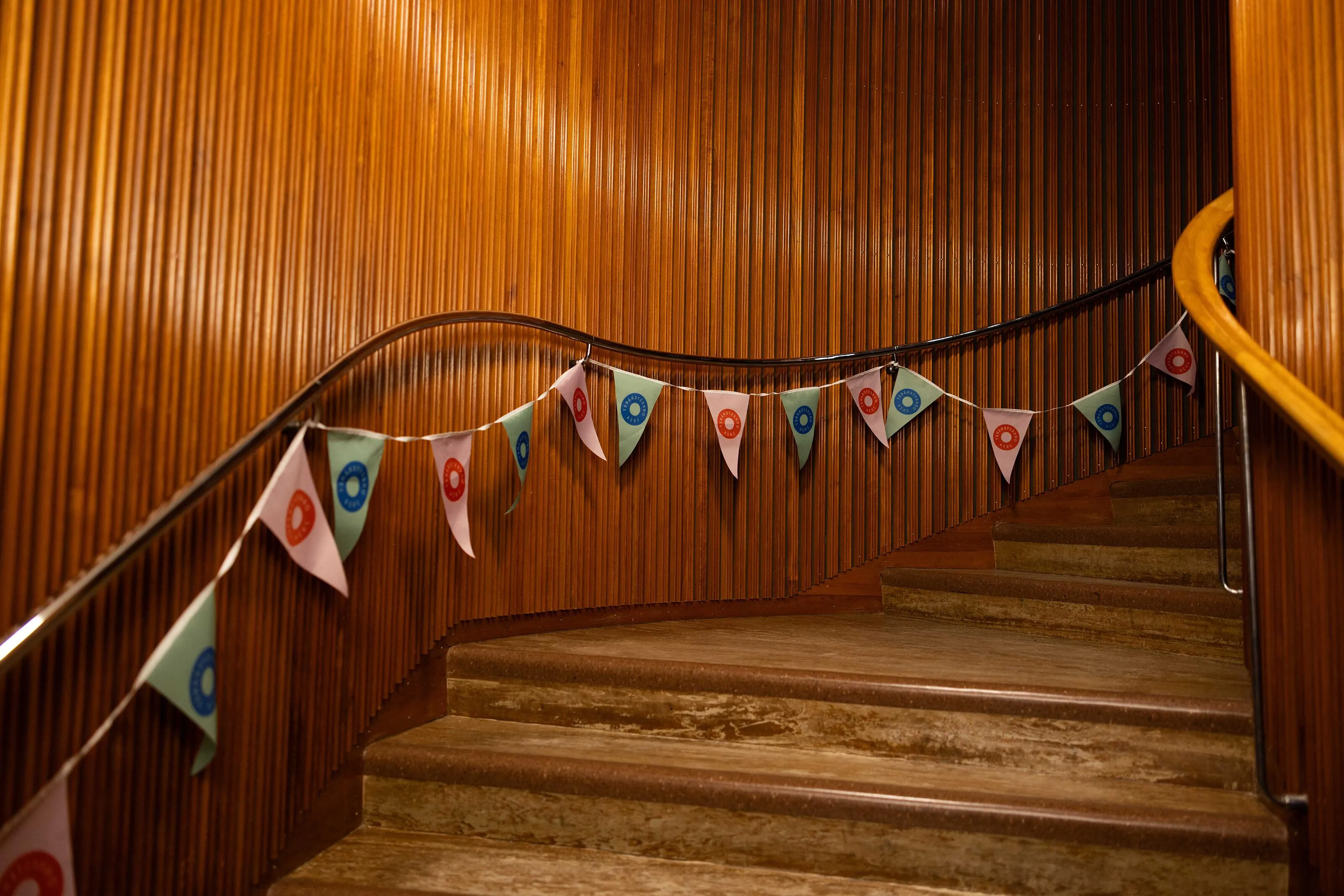 String of triangular flags decorated with Orkesteråret 2024 logos hanging along a curved wooden staircase, turning a concert venue into a festive celebration space.