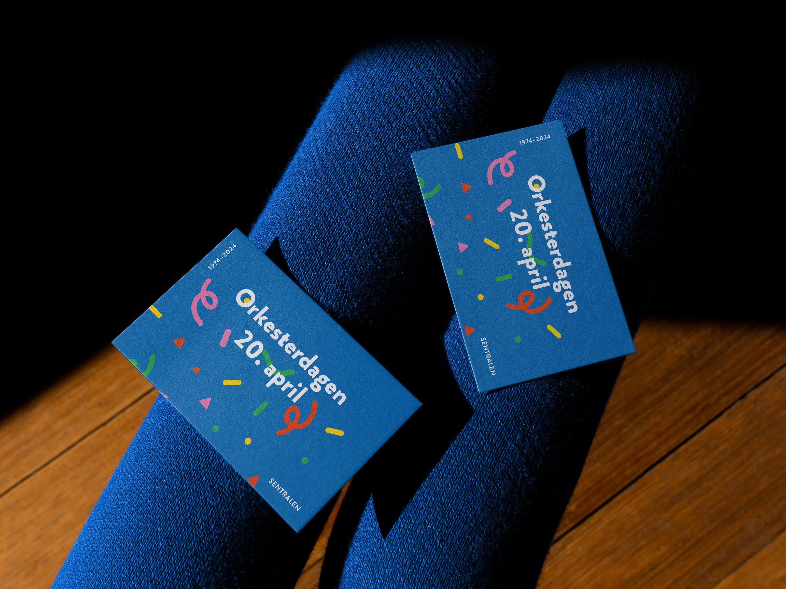 Two blue Orkesterdagen business cards with colourful confetti graphics lying on dark blue fabric in sunlight, promoting the national orchestra celebration day.