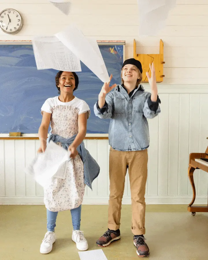 Two smiling school pupils in front of a blackboard throwing sheet music into the air, symbolising the joy and freedom of playing in an orchestra.