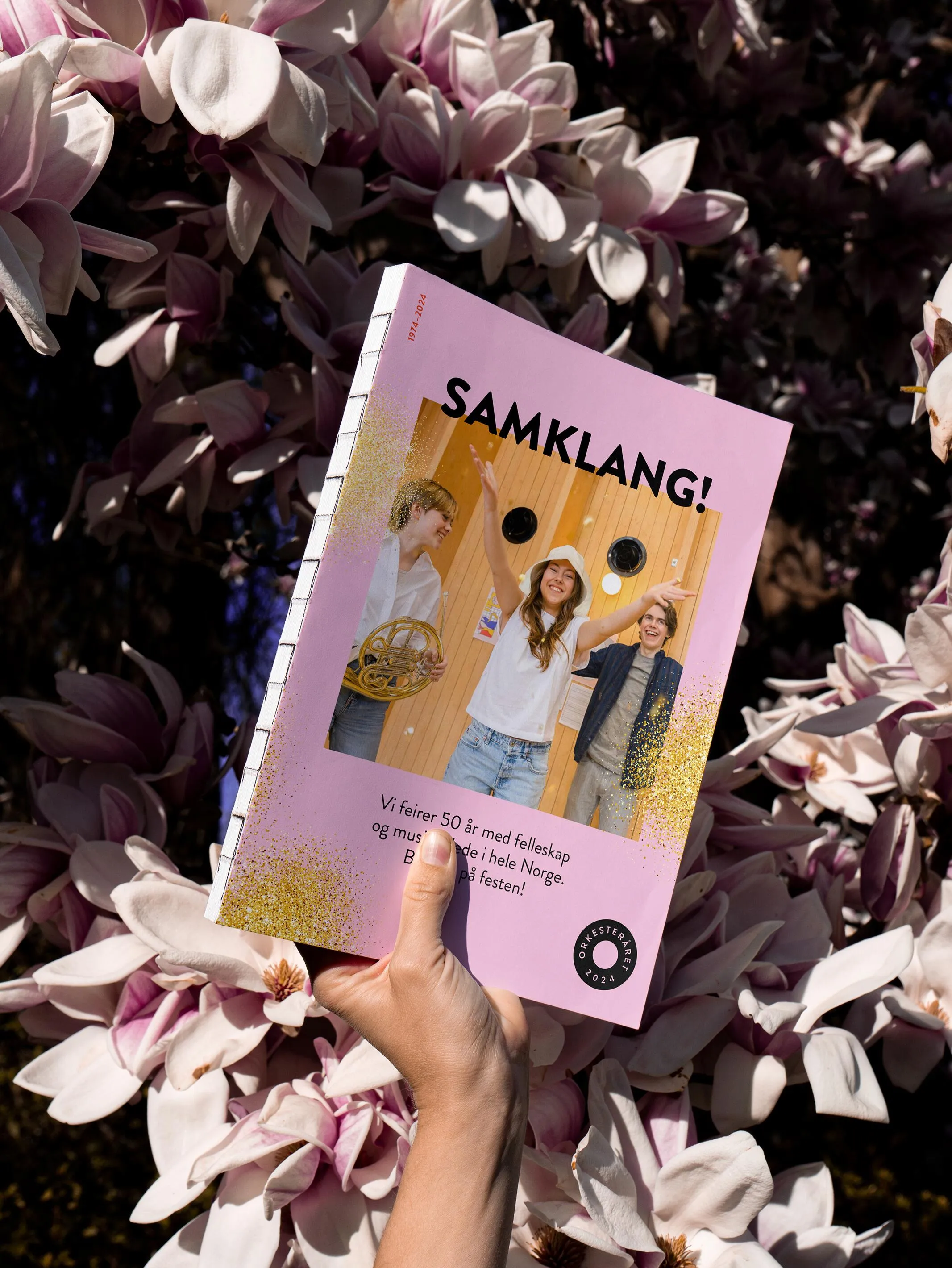 Hand holding a "Samklang!" programme booklet with Orkesteråret branding in front of blooming magnolia flowers, linking the visual identity to a festive spring setting.