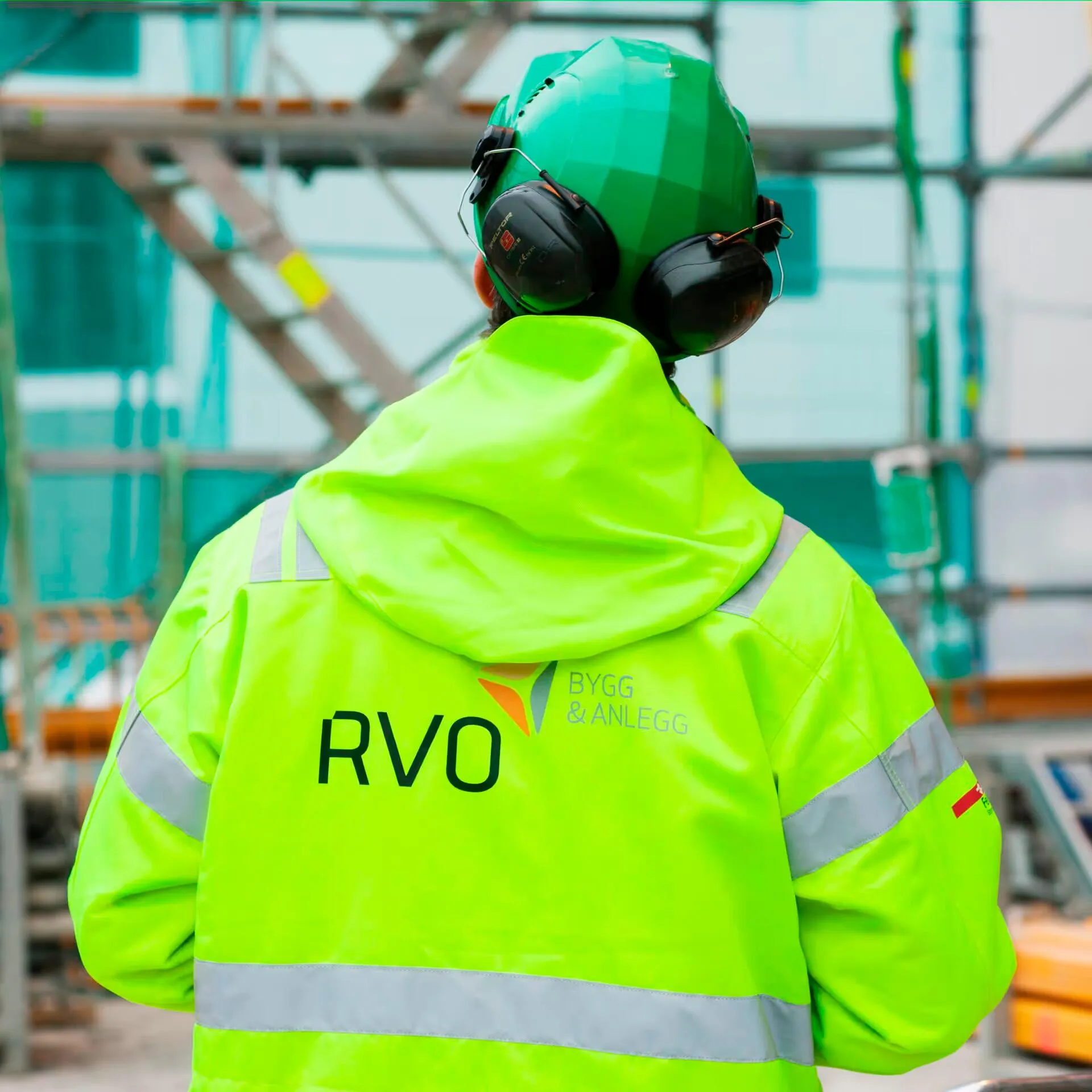 Construction worker wearing a green helmet, black ear protection, and a bright yellow high-visibility jacket with RVO and Bygg & Anlegg logos on the back.