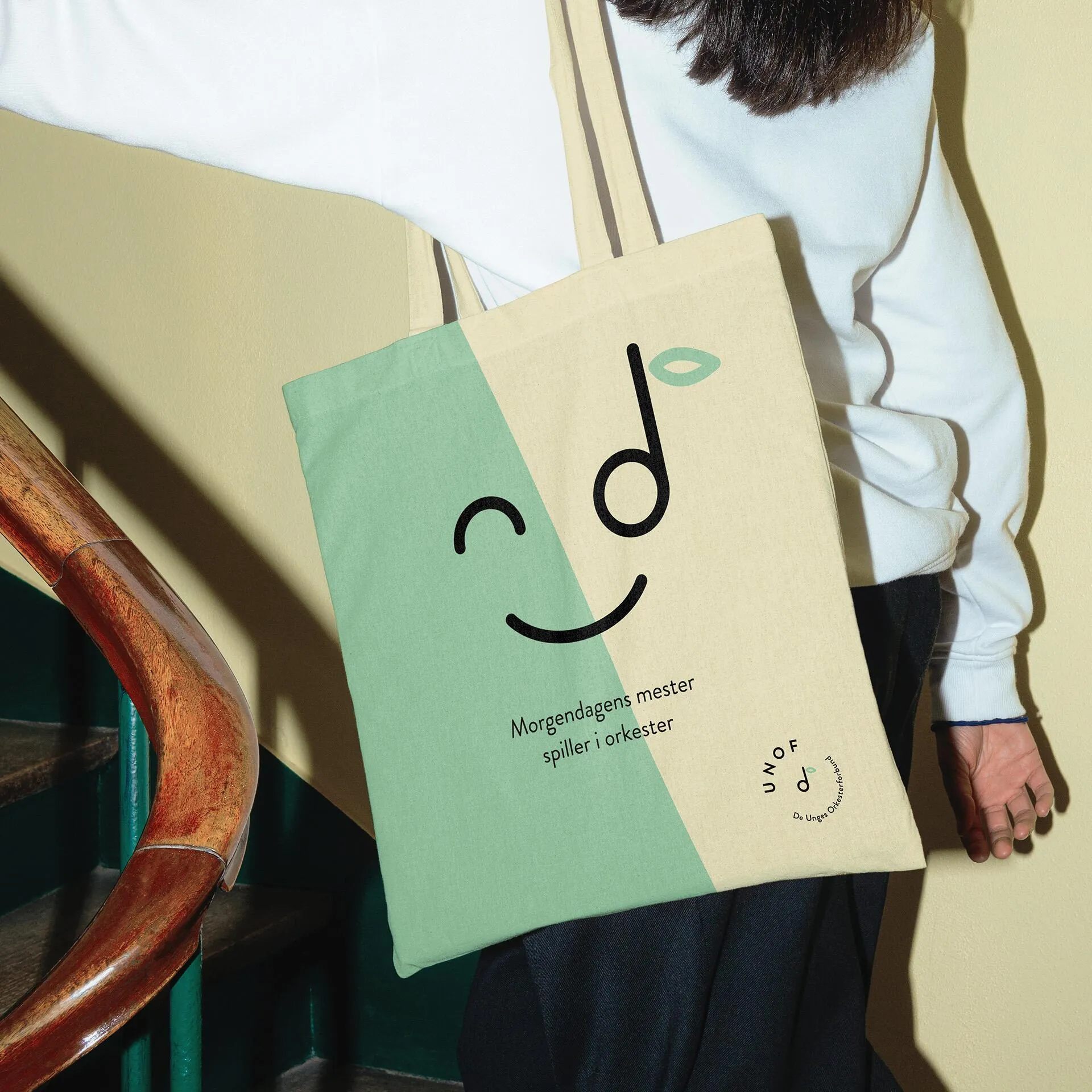 Person wearing a white sweater carrying a two-toned green and beige tote bag with a smiling face made of musical notes and the text 'Morgendagens mester spiller i orkester'.