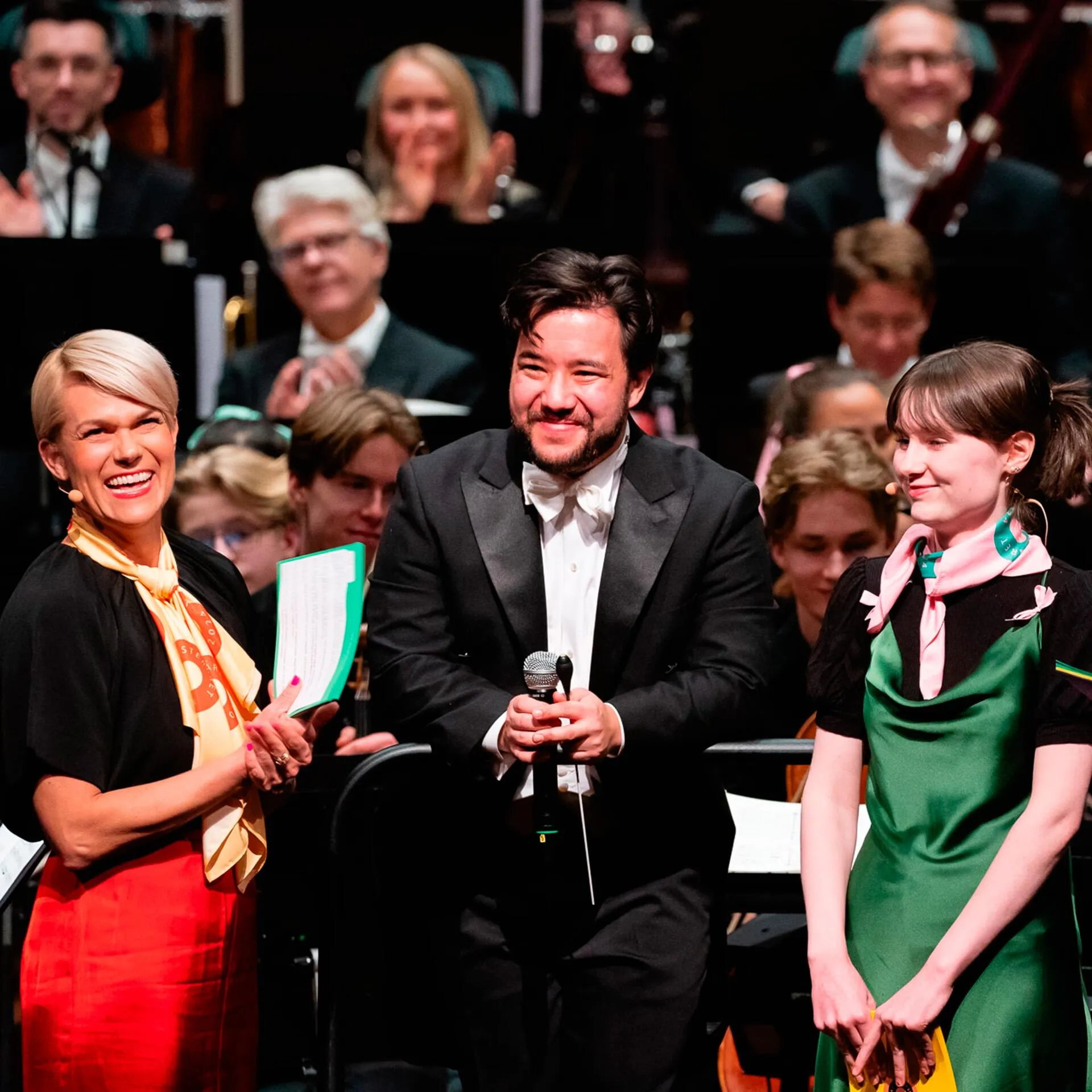 Three smiling people on stage in front of an orchestra, one holding a microphone and two holding papers.