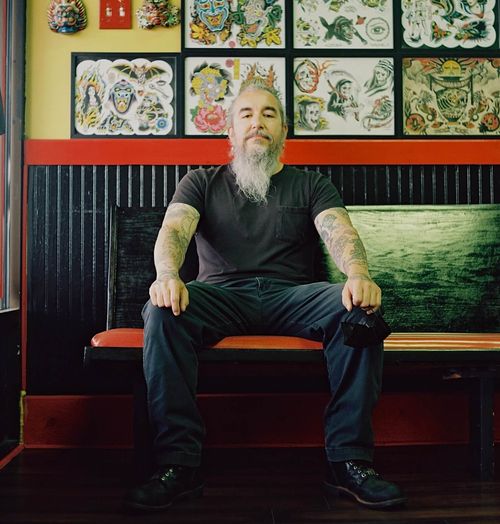JP Morin, of Saints and Sinners Tattoo, sitting in front of a wall covered in framed flash art drawings.